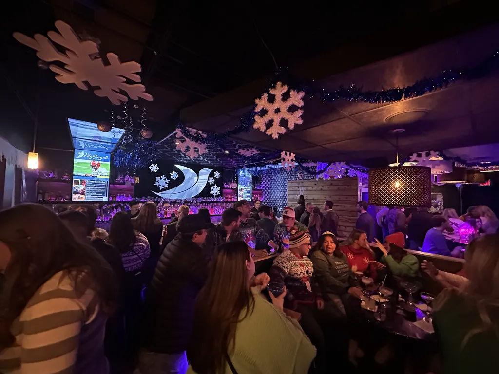 A festive crowd in holiday sweaters beneath hanging snowflakes and purple lights